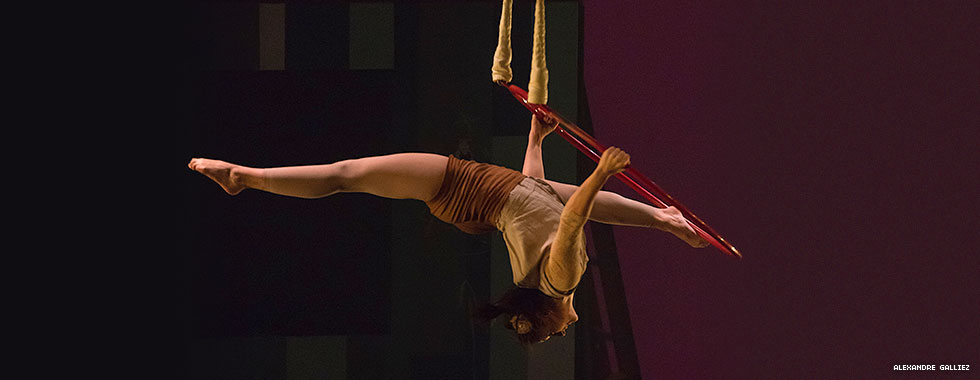 An acrobat outstretches her legs in opposite directions and bends her head and neck back while holding on to a hula hoop that's suspended from ropes.