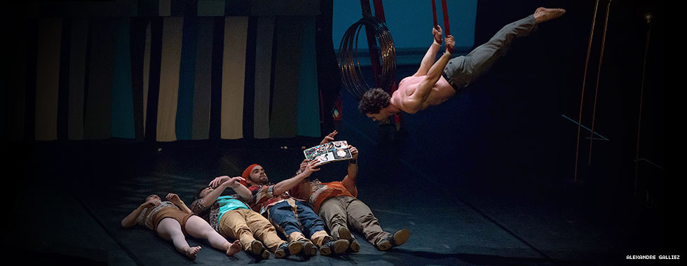 An acrobat hanging by his wrists from two suspended ropes swings low over four performers laying on the stage.