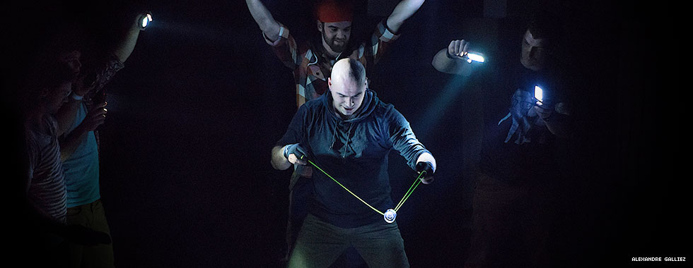 Circus performers hold flashlights on a performer who plays with a yo-yo.