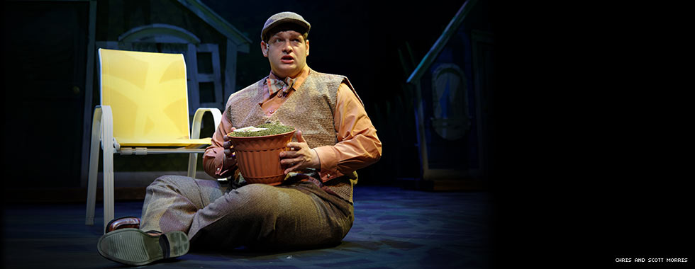 An actor wearing a vest, pressed slacks, and a beret sits next to a lawn chair and holds a flower pot.