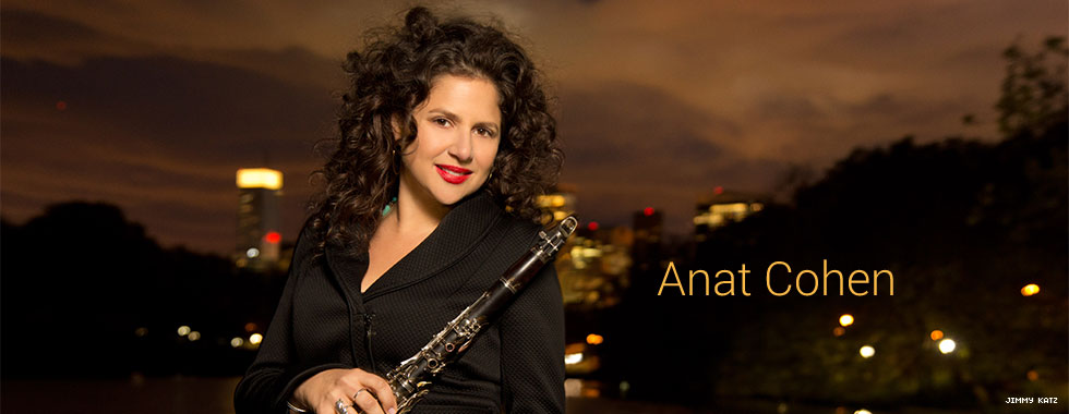Anat Cohen, with a nighttime cityscape in the background, holds her clarinet with her right hand, leaning it on her left shoulder, while she smiles for the camera.