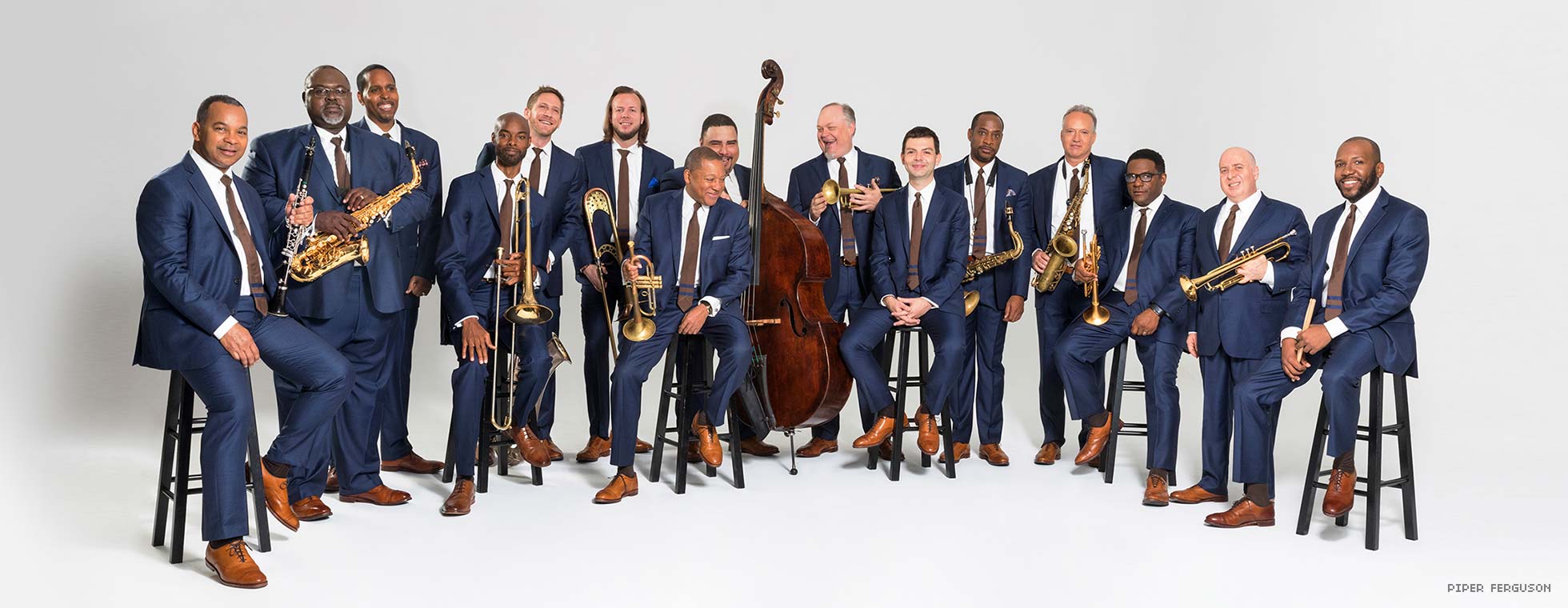 15 musicians holding their instruments and wearing suits, stand in a row with Wynton Marsalis at the center.