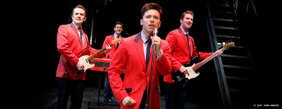 The Four Seasons, dressed in matching suit jackets, each pose with their instrument while one actor stands in the front at a microphone.
