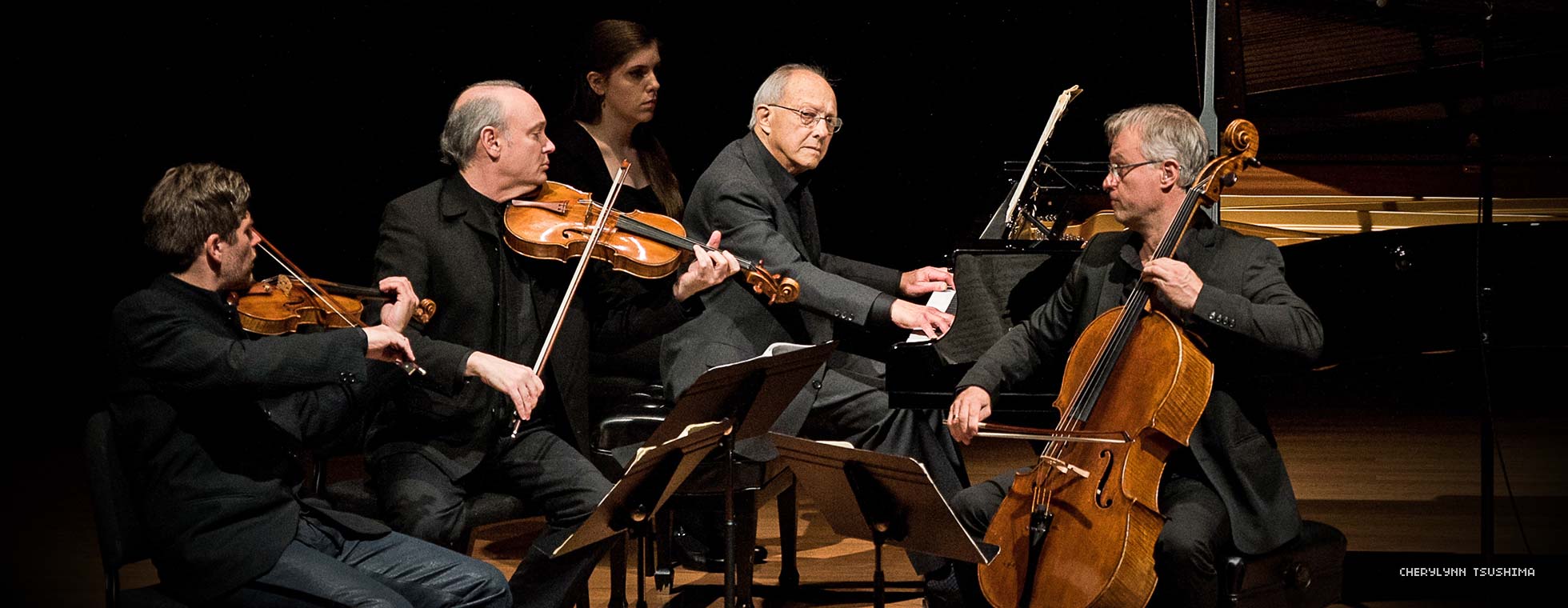 Various formally dressed string musicians are seated around a pianist who performs while he looks away from his instrument.
