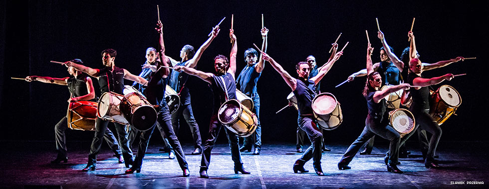 Twelve musicians each stand with their left arm raised and holding a drumstick while their drums hang from a strap around their shoulders.