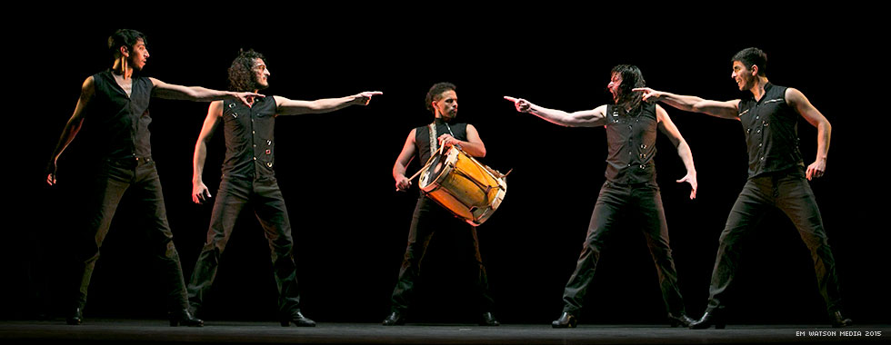 Four musicians point at a percussionist standing in the middle of them while he plays his drum.