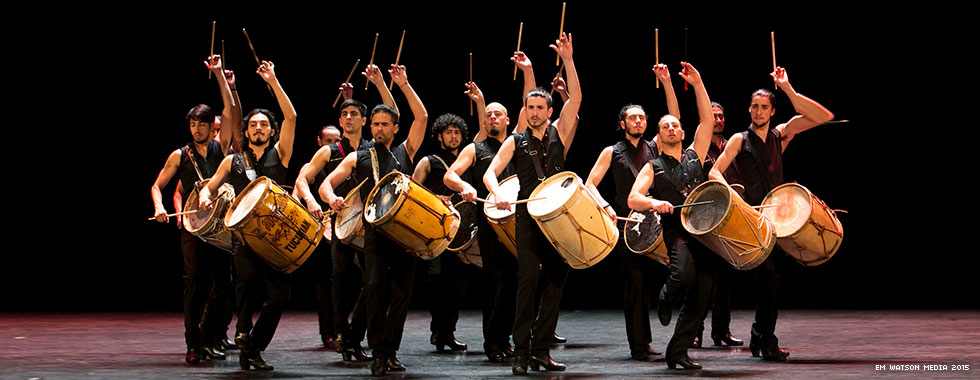 A group of musicians with drums strapped to their shoulders hold their arms above their heads or straight in front of them while they stand in a circle and look at the audience.