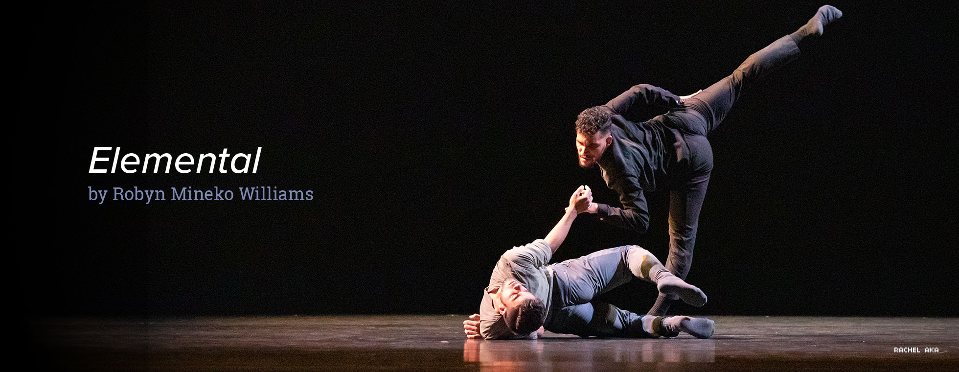 In a scene from “Elemental,” by Robyn Mineko Williams, a dancer lays on his side on the ground while another dancer bends over at the waist and points his right leg up and their hands meet. 