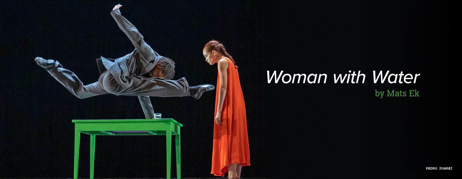 In a scene from “Woman with Water,” by Mats Ek, a dancer using one hand to hold himself above and parallel to a table stretches both legs in opposite directions while a woman stands and watches. 