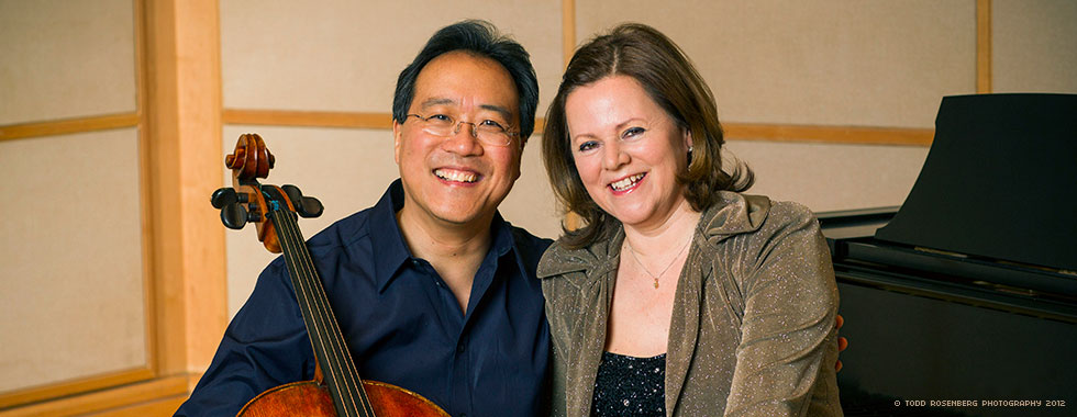 Yo-Yo Ma smiles while cradling his cello in his right arm and his left arm postured around a smiling Kathryn Stott.