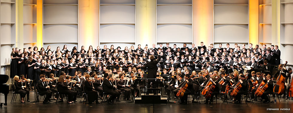 An orchestra performs in a semi circle around a conductor while members of a large chorus, each holding a songbook, stand on risers behind the musicians.