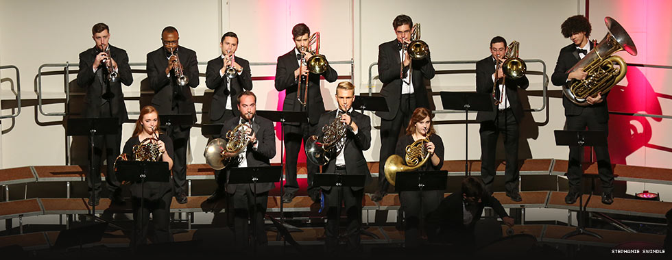 Eleven musicians playing a variety of brass instruments stand at their music stands in two rows.