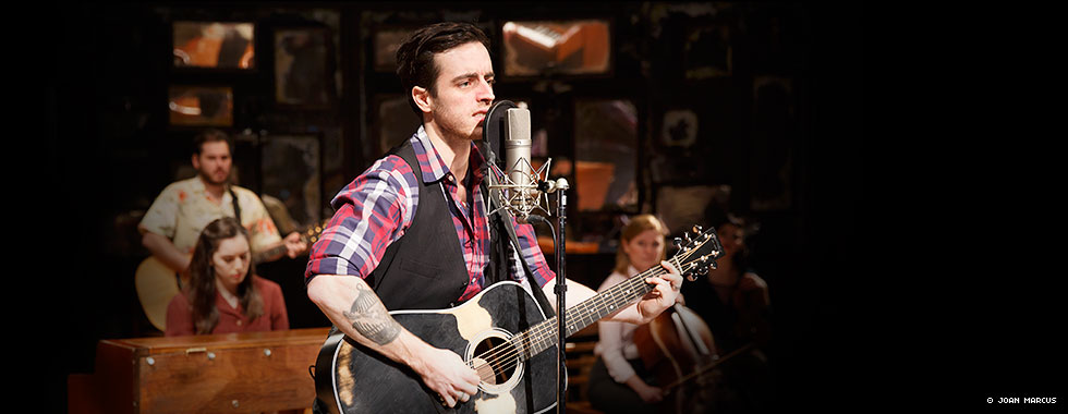 "Guy," the main character in "Once," sings into a microphone and plays guitar while musicians and members of the cast in the background assist in the musical performance.
