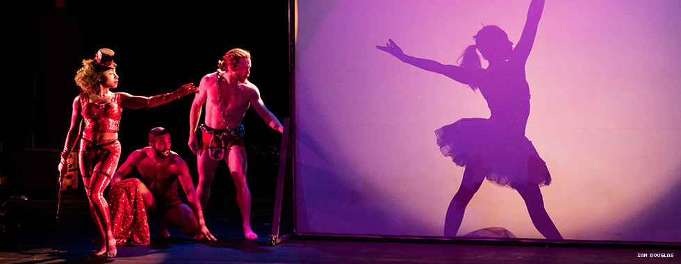 Two shirtless male dancers and a female dancer dressed in leggings and asporty bodice watch a dancer dressed in a tutu posing behind a screen in Pilobolus' "Shadowland."