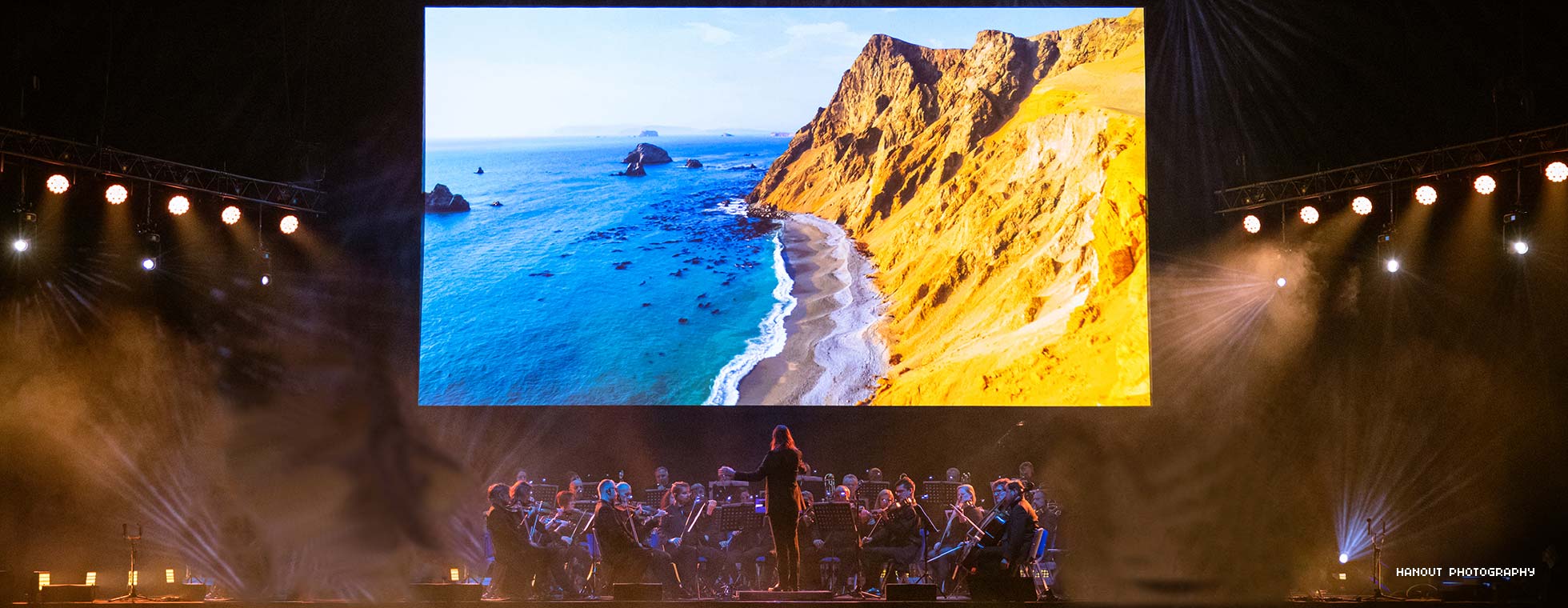 A man with outstretched arms conducts an orchestra seated below a video depicting a bird’s eye view of waves gently rolling into the side of a majestic cliff. 