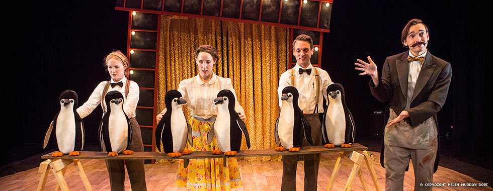 A man wearing a suit jacket gestures to three actors each standing in front of two stuffed penguins.