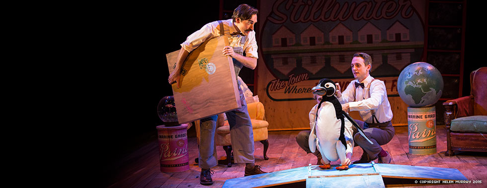 A man stands and holds a wooden box while another man crouches next to a stuffed penguin.