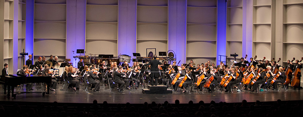 A conductor stands on a pedestal in front of the orchestra. 