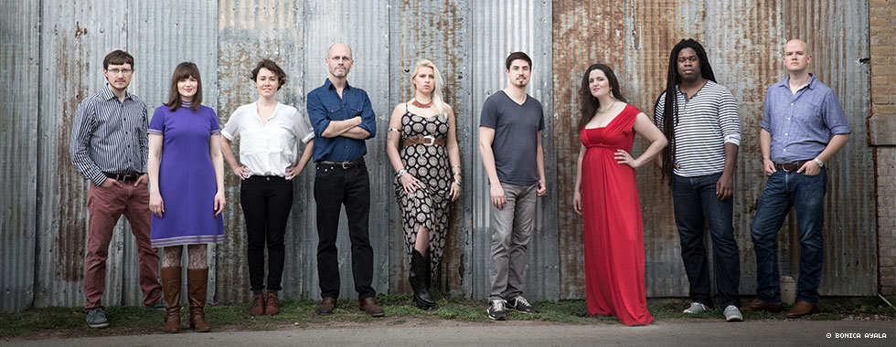 All nine members of Roomful of Teeth—eight singers and the artistic director—stand in front of a corrugated metal wall and smile for the camera.
