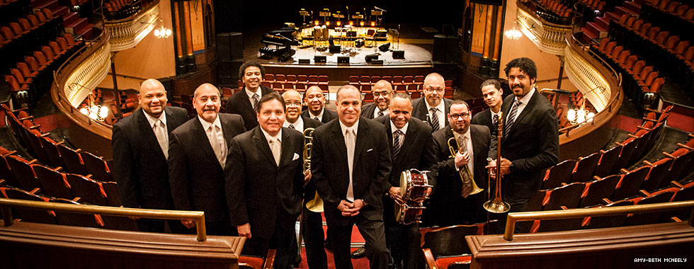 13 ensemble musicians, some of whom hold their brass or percussion instruments, stand among seats in the balcony level of an auditorium with the stage visible in the background.
