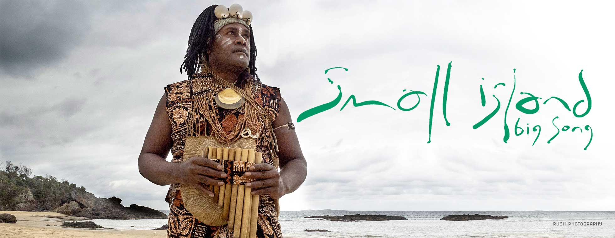 A musician wearing indigenous clothing holds a wind instrument and looks out at the ocean while standing on a tropical beach. 