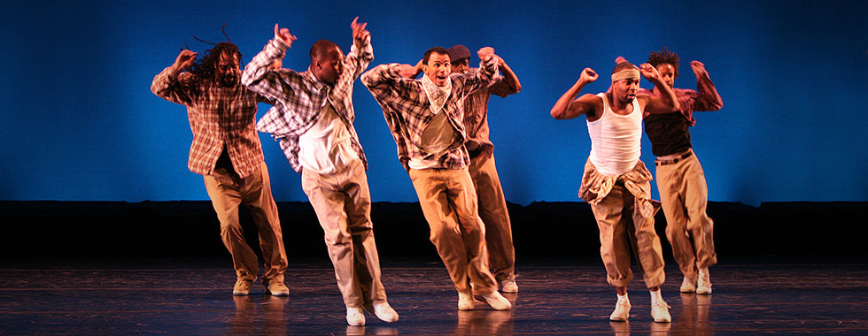 Six dancers wearing baggy pants and tank tops or plaid button-down shirts jump with both feet into a slanted position while they hold their arms up bent at the elbow.