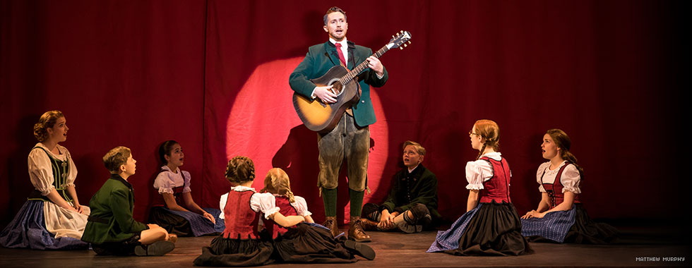 Captain von Trapp stands proudly as he plays his guitar while singing among Maria and his children who are sitting in a circle around him.