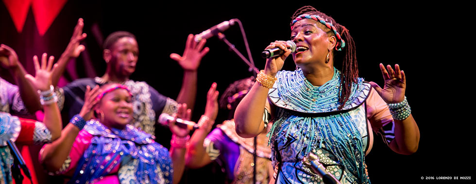 A woman wearing a braided headband, bracelets, and a dress adorned with beads sings while a members of a chorus in the background raise up their arms.