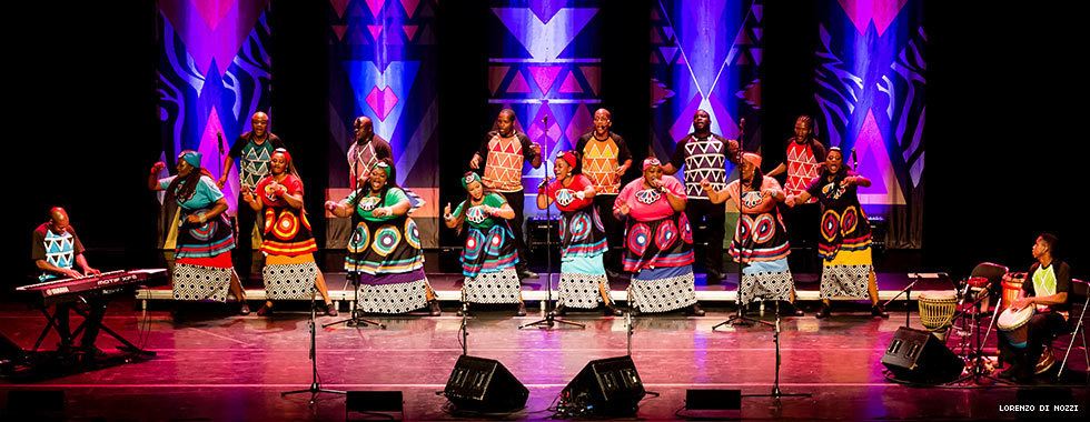 A chorus of men and women wearing costumes featuring similar designs sing and dance while a keyboardist and congo drummer perform on opposite ends of the group.