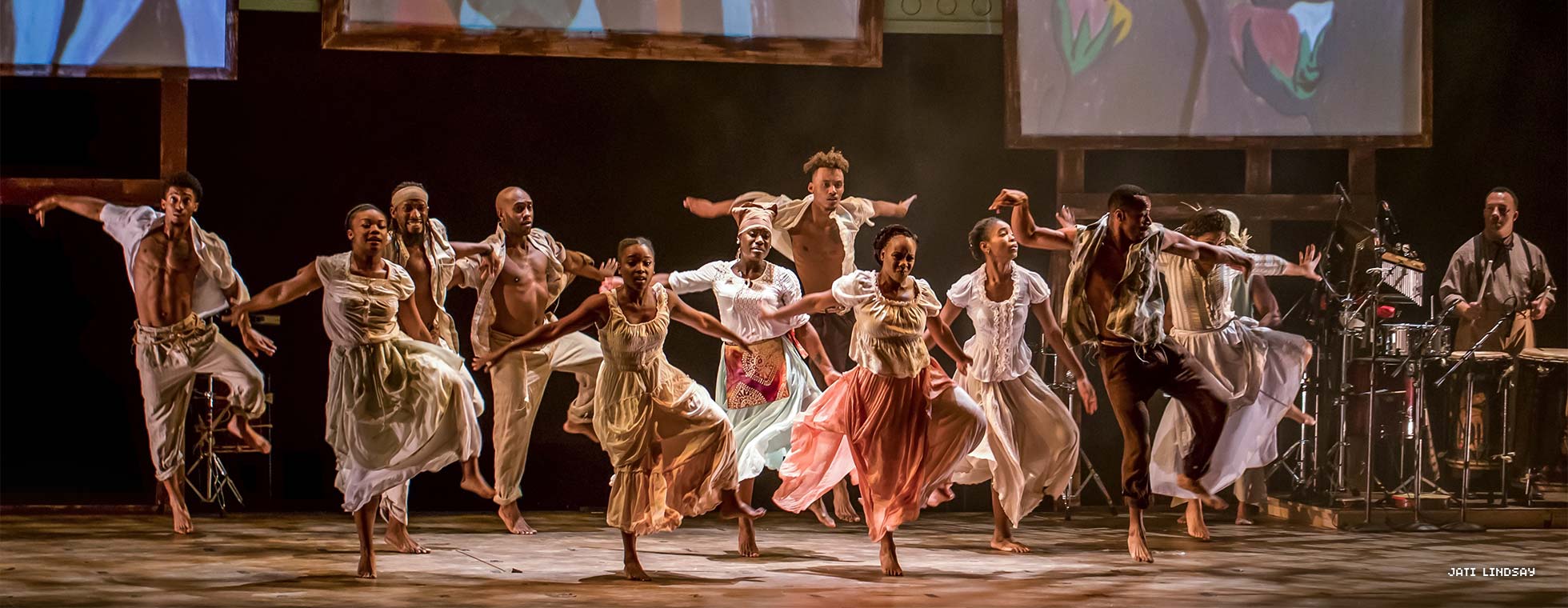 A group of Black dancers wearing loose clothing step dance in unison on a stage. 