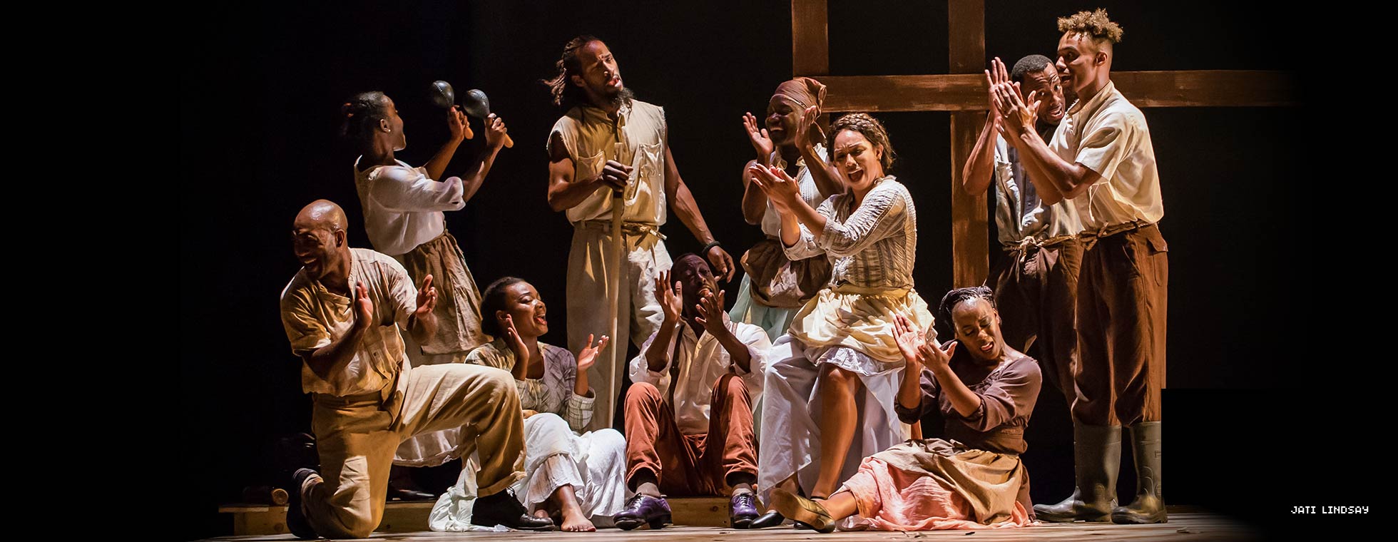 Black dance artists cluster together and clap their hands and sing. 