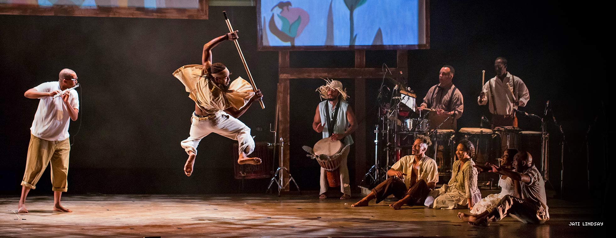 An artist uses a stick to propel himself into a dance flip while a small group of dancers watch. 