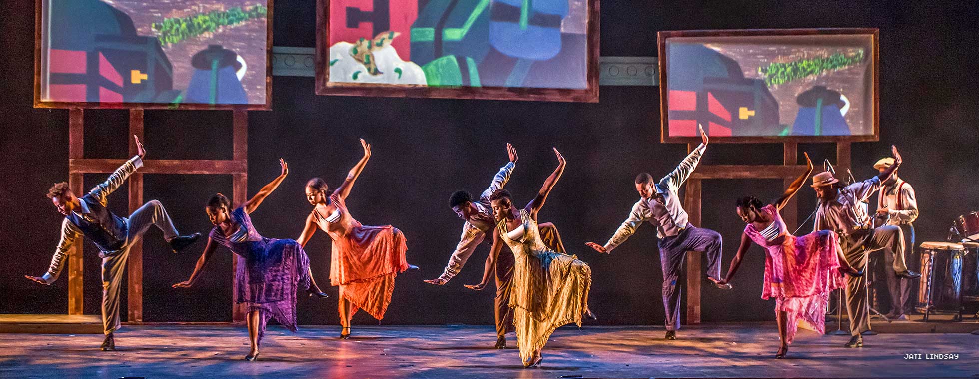 Five Black dancers wearing loose clothing pose in unison with both arms outstretched. Paintings are projected behind them. 