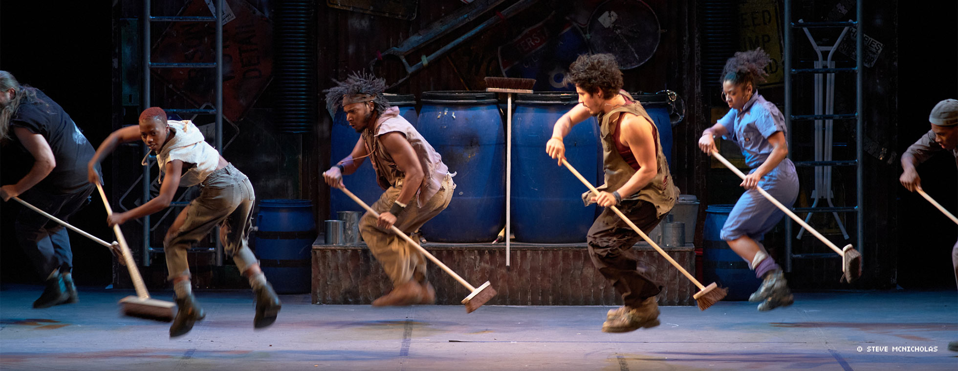 Men of color facing the same direction hold utility brooms in a sweeping motion while they jump in mid-air.