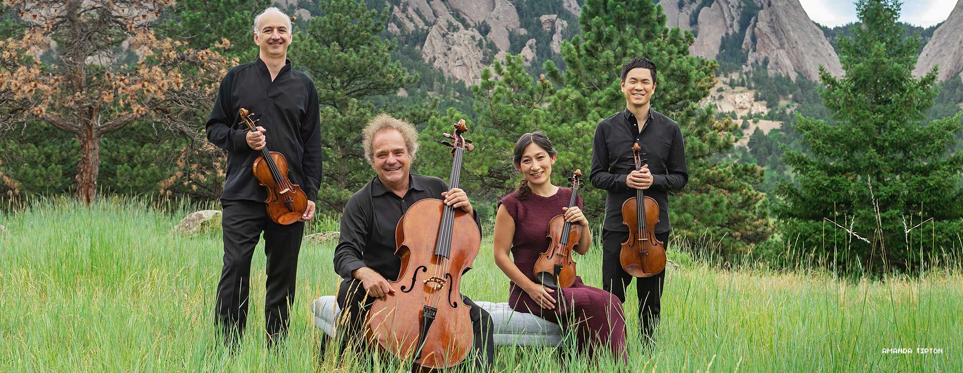Four musicians posed in an idyllic mountain pasture hold their stringed instruments.