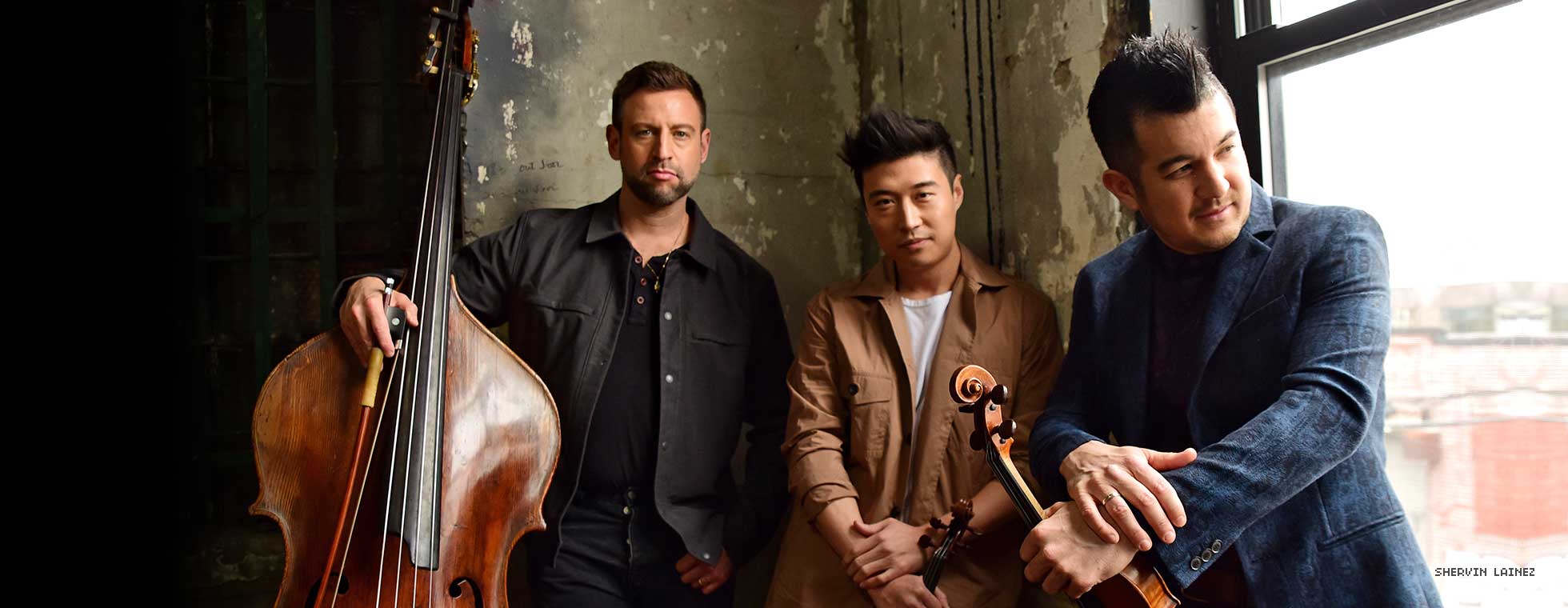 Three chic but casually dressed men hold their string instruments while standing in front of a window. 