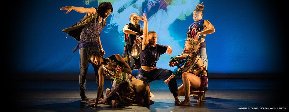 A group of dancers are shown in various poses with arms stretched or legs and knees bent on a stage in front of an image projected on a screen.