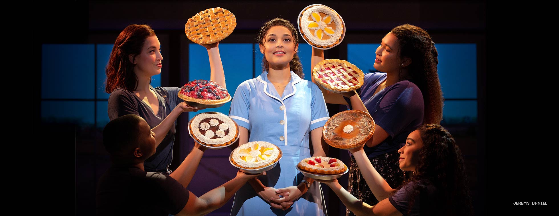 A woman dressed in a vintage diner-style shirt dress server’s uniform stands and is framed by four women holding various pies.