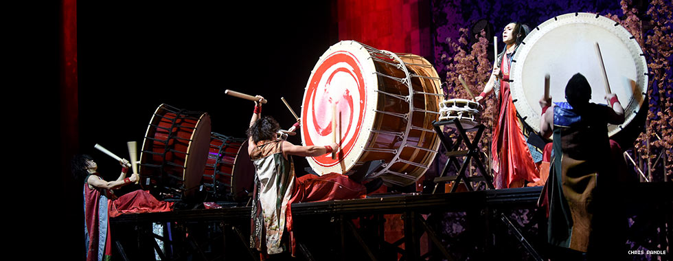 Three musicians play large drums set on their side on a platform while a fourth performer stands on the platform and plays a smaller drum.