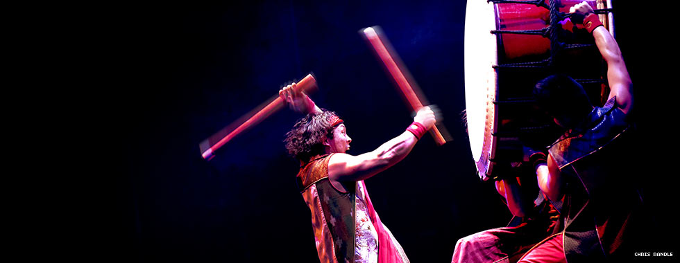 A percussionist with two drumsticks bangs on a large drum held upright by two men.