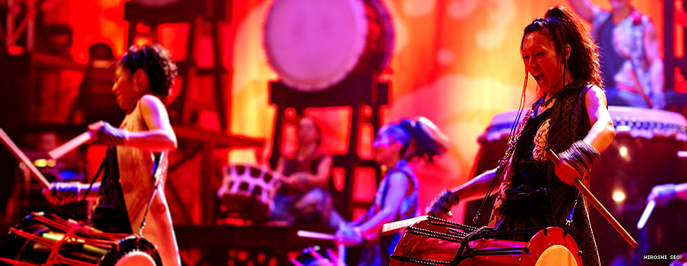 A drummer yells out while playing a drum suspended by a harness from her shoulder.