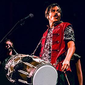 A man with a mustache wears a large two-sided drum around his neck.