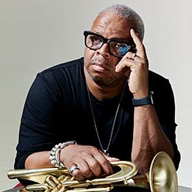 A Black man wearing fashion-forward eyeglasses leans his head against one arm while he holds a trumpet with the other.