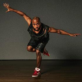 A Black man wears athletic-inspired gear while he lunges down and spreads his arms out on each side.