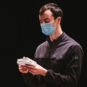 A man wears a buttoned-up shirt and a face mask while he looks down at index cards he’s holding with both hands. 