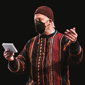 A man wearing a knit cap, face mask and striped 80-style sweater extends his left arm out while he looks down at index cards in his right hand.
