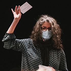 A person with long hair and eyeglasses and wearing a face mask holds an index card slightly over her head.