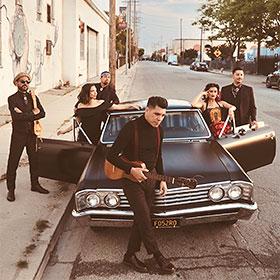 Monochromatically dressed artists stand around a modified antique car parked on a city street.