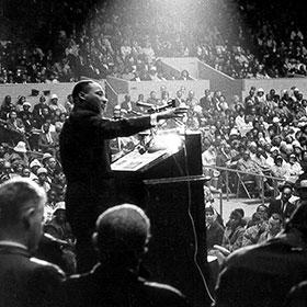 An historic photo shows Dr. Martin Luther King Jr. Standing at a lectern and speaking to a vast audience while a spotlight shines on him from above.