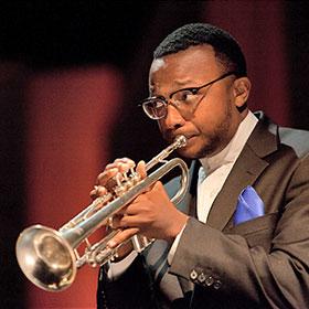 A Black man wears eyeglasses and a stylish suit as he plays a trumpet.